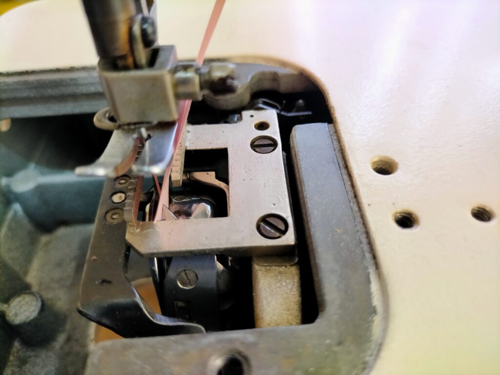 brother TA3-B627 職業用ミシンの釜調整実施中の実写。針と釜の剣先の間にピンク色の付箋紙(ノギスで測定した約0.08mm)を挟んで隙間を0.04〜0.1mmに調整している状態。付箋紙をシムゲージ代わりに使う実践的な調整方法