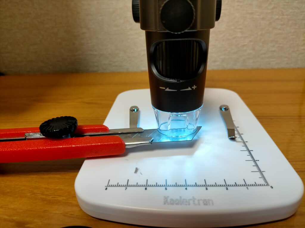 Koolertron デジタル顕微鏡でカッターの刃を高倍率で観察した画像①。対物レンズを刃に最大限近づけた高倍率撮影。刃こぼれと、ガムテープを切った際に付着した粘着物が確認できる。肉眼では見えない細部まで観察可能な実例