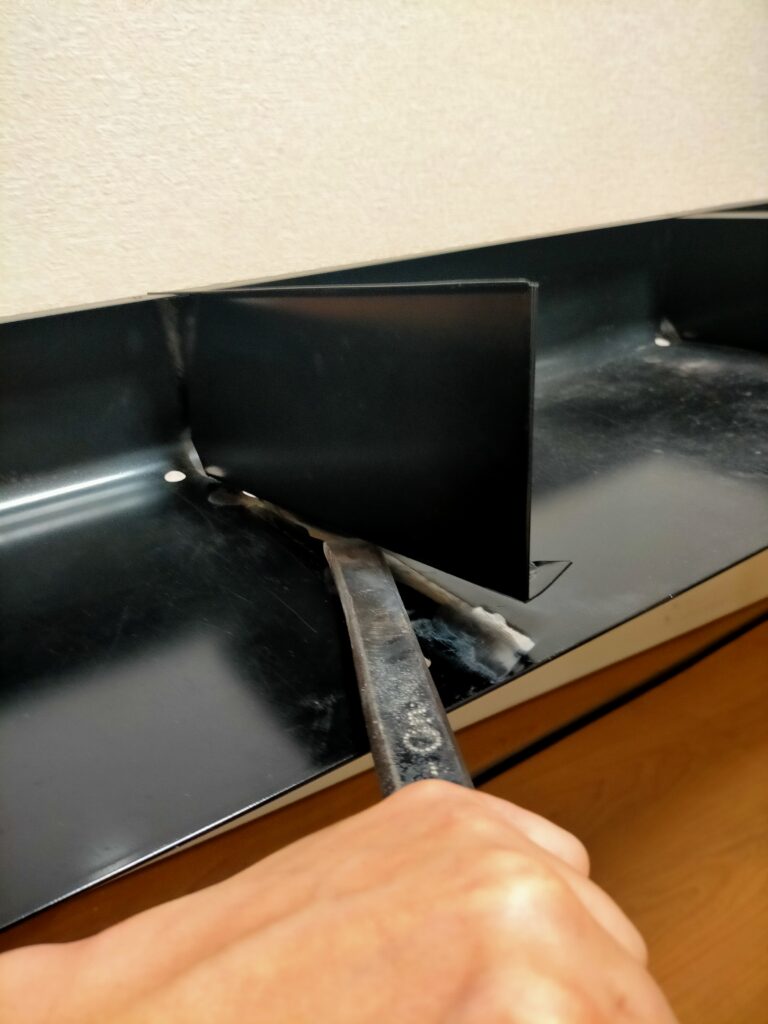 サンワ 100-KB004 キーボードスライダー取り付け作業Step2。手製のタガネ（2トン車のリーフスプリングを加工した愛用工具）で机の鉄板間仕切りのスポット溶接4カ所を切り離している状態。キーボードスライダーのクランプ取り付けを妨げる間仕切り撤去DIY