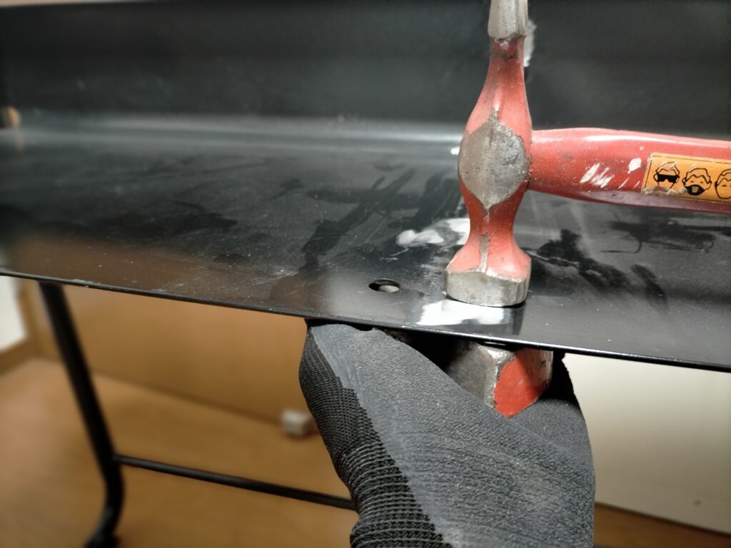 サンワ 100-KB004 取り付け作業Step4・板金仕上げ。タガネ作業で曲がってしまった机の鉄板を板金ハンマーで叩いて均している状態。グラインダーでバリ取り後・シャーシブラック塗装前の板金整形作業