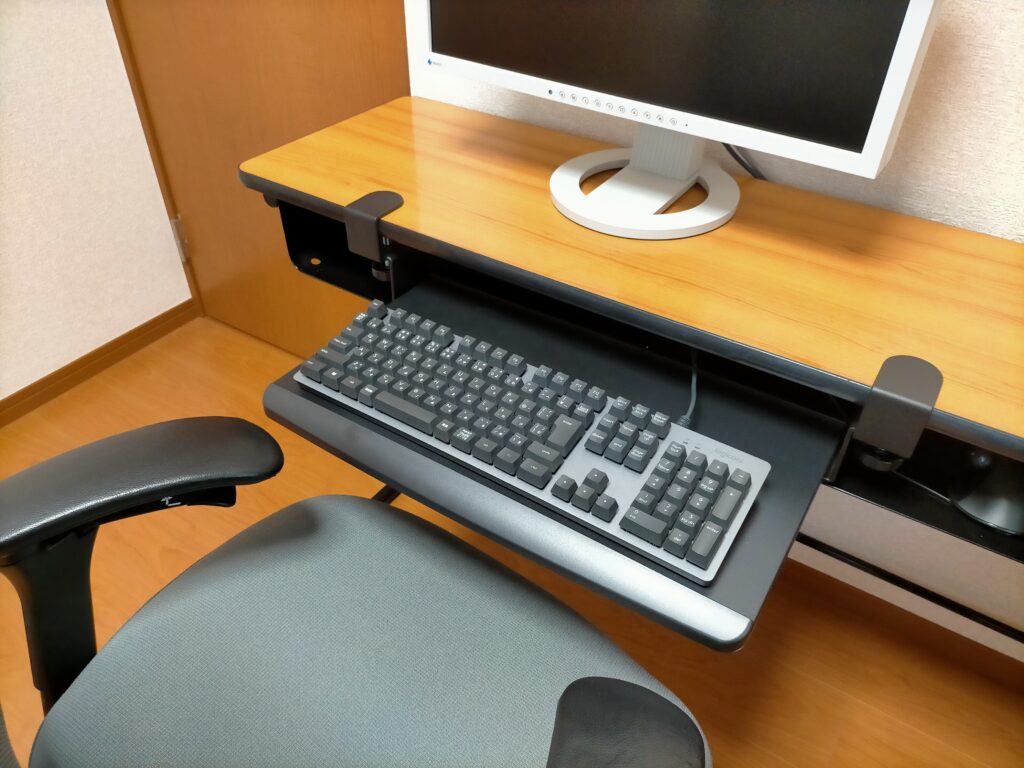 サンワ 100-KB004 キーボードスライダー取り付け完了（After）。幅51cmのスライダーがクランプで机天板下に固定された状態。ロジクール K840 フルキーボード（テンキー付き）を載せた使用状態。オカムラ コンテッサの肘置き高さとキーボード位置が一致してタイピング姿勢が改善