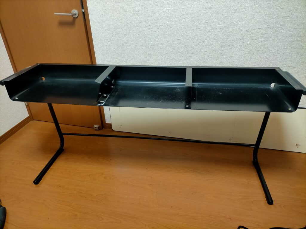 サンワ 100-KB004 キーボードスライダー取り付け作業Step1。机のビスを外して天板を取り外した状態。天板下の収納スペースにあるキーボードスライダーのクランプ取り付けを妨げる鉄板の間仕切りが露出。4カ所スポット溶接された間仕切りの撤去前