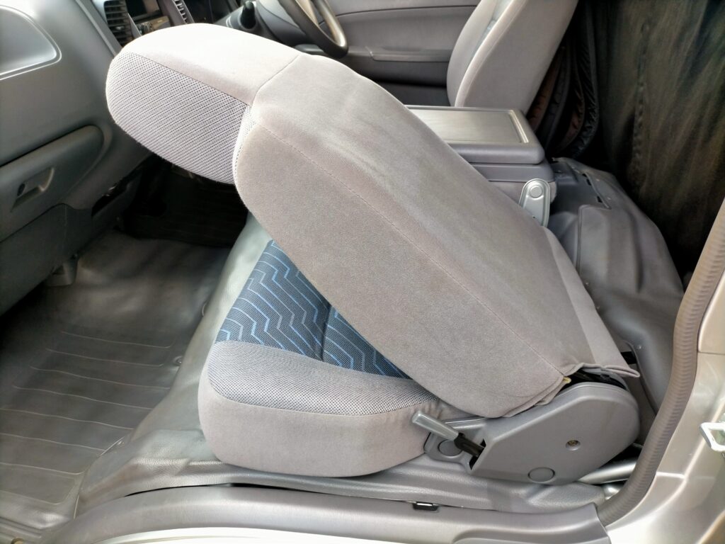 E25キャラバン／コモのエンジンアクセス手順ステップ2・助手席の背もたれを前に倒した状態。キャブオーバー車特有の構造でシートの下にエンジンが配置されており、背もたれを前倒しすることが座席跳ね上げの前準備。この後座席ごと持ち上げてKA24DEエンジンルームを露出させる