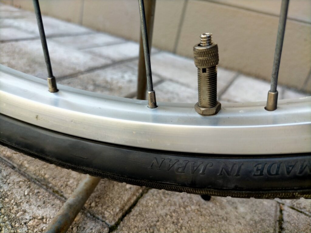 自転車パンク修理ステップ1・虫ゴム交換の手順2。英式バルブのトップナット（指で回せるローレットナット）を手で緩めて外している状態。工具不要で手回しできる。次にプランジャーを引き抜く前の工程