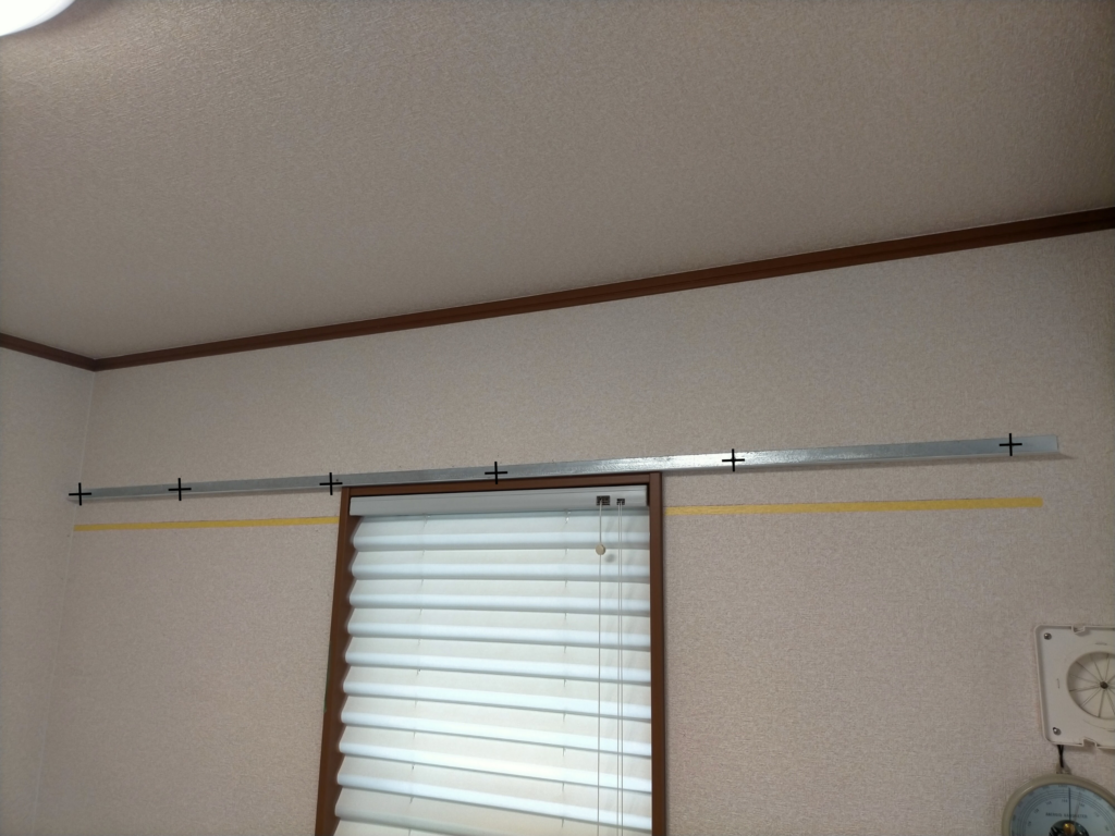 フローティングシェルフDIY・アングルを実際の壁取り付け位置に仮置きして穴あけ位置を現物合わせでケガく工程の図解。仮置きによる現物合わせで穴位置のズレを最小化。「×」印が正確な穴位置マーキングのポイントを示す