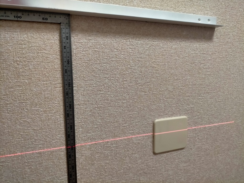 フローティングシェルフDIY・レーザー墨出し器のグリーンラインを基準に棚受けアングルの仮置き・取り付け位置をマーキングしている工程。窓枠両脇の柱と450mm間隔の間柱位置に合わせてアングル4本を配置。下地探し「どこ太」との併用で強固固定を実現