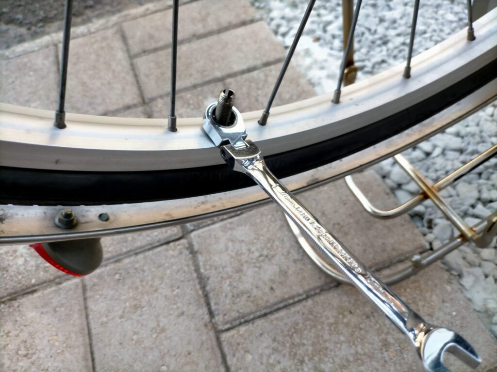 自転車パンク修理ステップ2・チューブ取り出し準備。バルブ根元のバルブ固定ナット（リムナット）を10mmメガネレンチで外している状態。工具が必要な唯一のナットで、これを外すことでバルブがリムから引き抜けるようになる