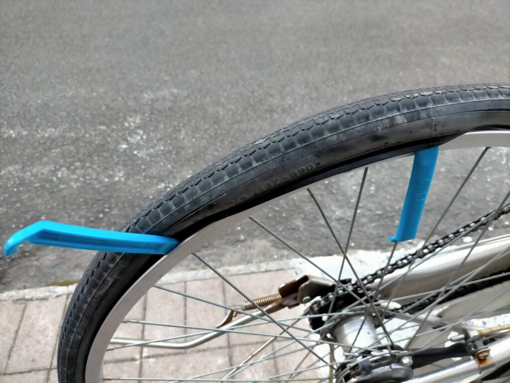 自転車パンク修理ステップ2・タイヤレバー2本を使ってタイヤビードをリムから外す手順。1本目のレバーをスポークに引っ掛けて固定し、2本目で10〜15cm隣を同様にめくる作業を繰り返す。自転車を逆さにしなくても作業可能