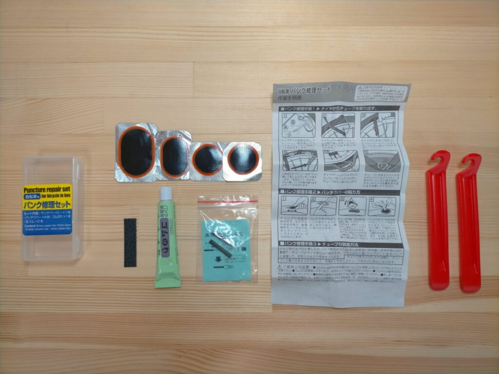 自転車チューブパンク修理に必要な工具・材料一式。パンク修理セット（パッチ・ゴムのり・紙やすり）、タイヤレバー2本、10mmメガネレンチまたはスパナ、バケツ（水張り用）、ゴムハンマー。100円ショップや修理セットで揃えられる