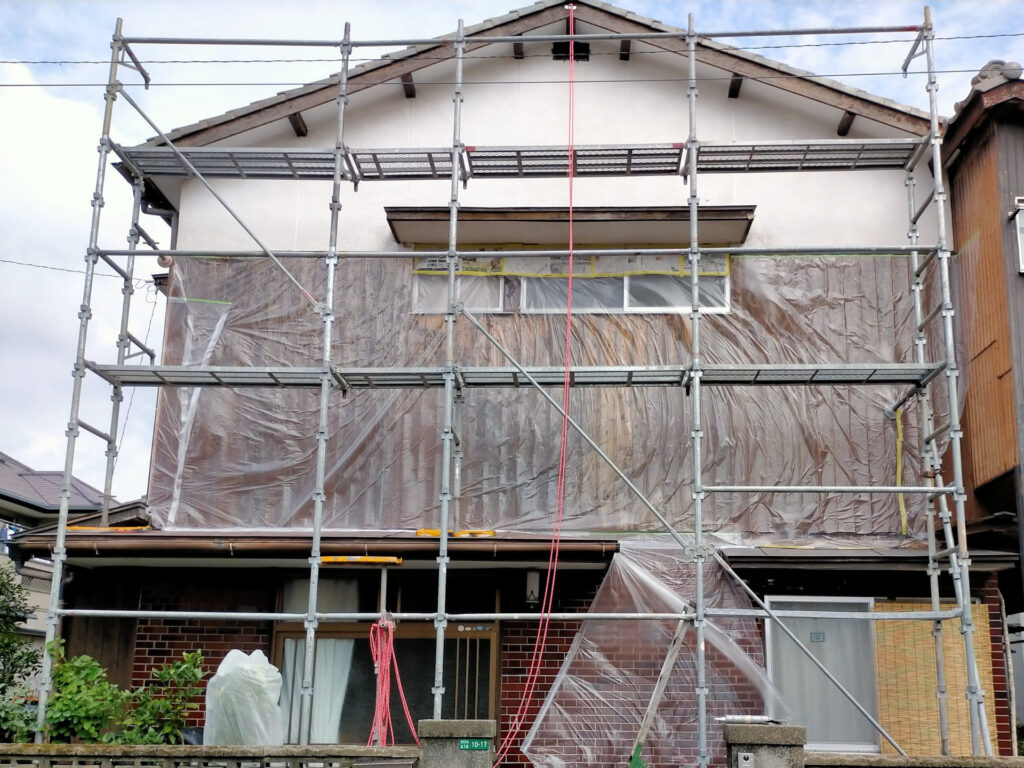 モルタル外壁塗装前の西面マスキング作業を完了した様子。マスキングテープ・養生テープ・養生マット・ポリマスカー・新聞紙で窓枠・基礎・設備配管など塗料が付着してはいけない箇所を養生した状態。足場設置済みの西面