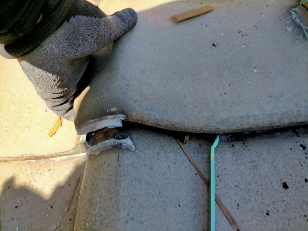 上段の瓦を少し持ち上げて白幡瓦用資材止G-6のフック部分を瓦に引っかけて差し込む取り付け工程。2カ所設置して角材(垂木)を渡すことで傾斜屋根上に水平な足場ベースが生まれる。瓦を傷めないDIY屋根作業手順