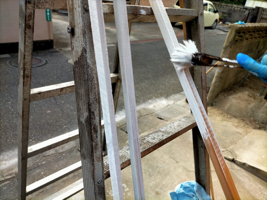 軒天ケイカル板固定用の杉角材（1980×15×15mm）を現場でカットする前に地上でカチオンシーラーと水性シリコン塗料で防腐塗装している工程。ケイカル板と同様に取り付け前に塗装することで高所作業の負担を軽減。塗装済みで切り出し後に現場固定