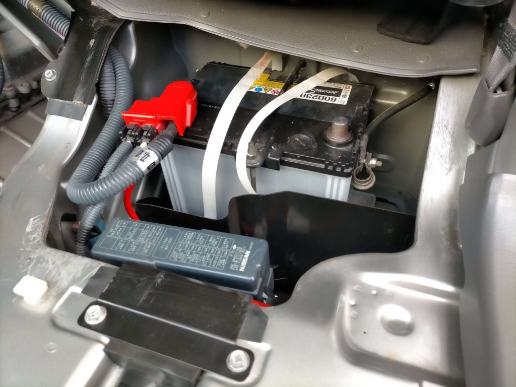 いすゞコモ（E25キャラバン）助手席下バッテリーのマイナス端子を10mmナットで取り外している作業。ETC2.0車載器DIY取り付けの最初の安全手順でパワーウィンドウ等の電装系トラブルを防ぐ