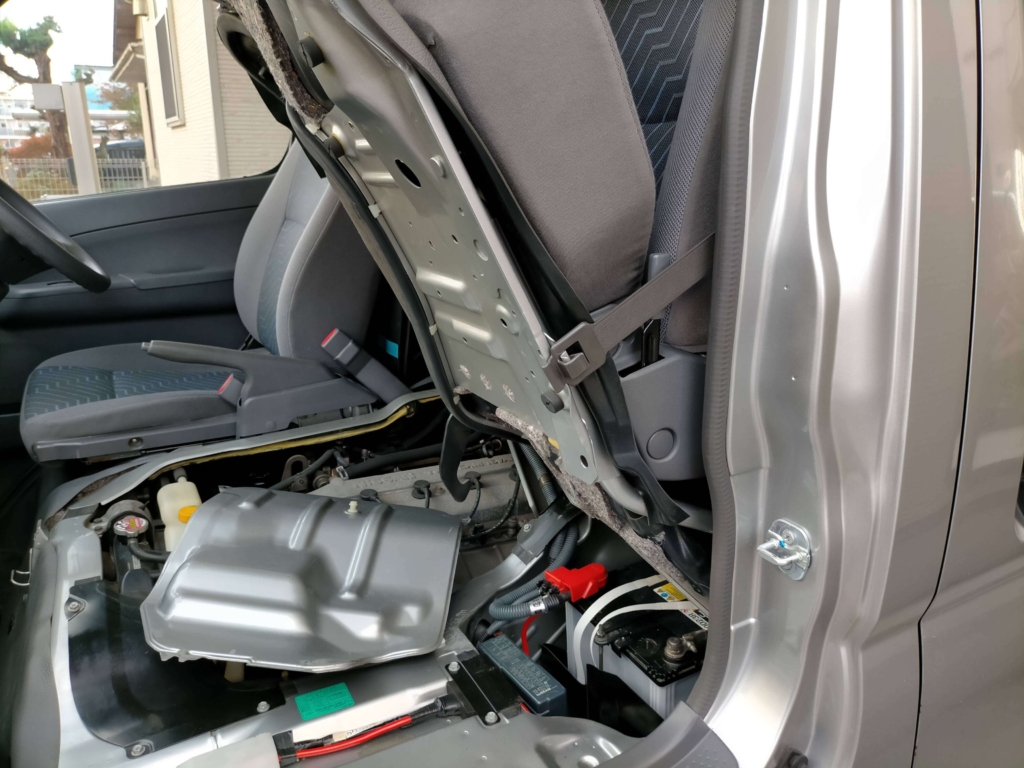 いすゞコモ（E25キャラバン）の助手席シート下に搭載されたバッテリーの位置。ETC取り付け作業開始前のマイナス端子取り外しのためのバッテリー確認。ショートや電装系トラブル防止のための基本作業