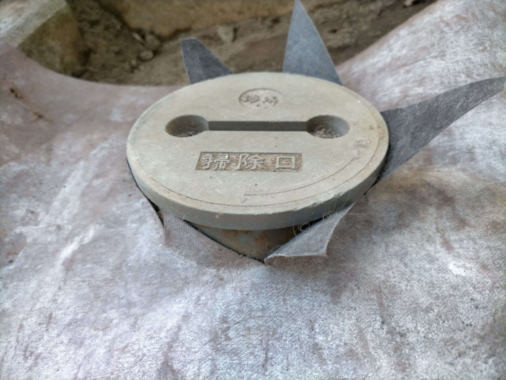 防草シートザバーン240をガス管・水道管・汚水桝部分に合わせてハサミ・カッターで切り込み加工を施している作業の様子。障害物周辺の防草シート施工の重要工程