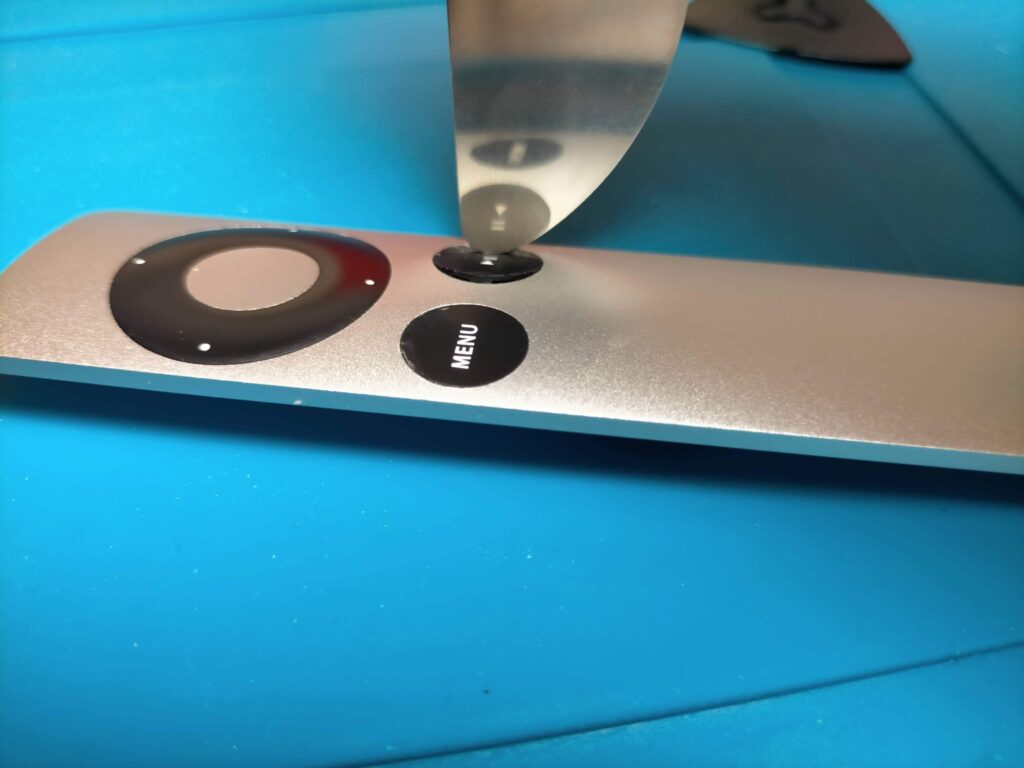 Apple Remoteの再生一時停止ボタンを金属製ヘラでこじって取り外そうとしている作業（ヘラを隙間に挿入している状態）