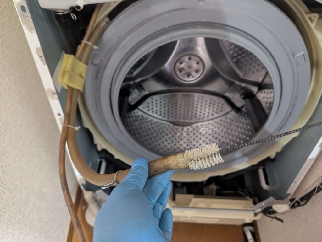 ドラム式洗濯機の循環ホース内をチューブブラシで清掃している状態。黒いワカメ内部診断Step5の確認箇所。柔軟剤・洗剤カスのヘドロが詰まって循環ポンプの圧送力が低下する原因。エアで吹いた後にヘドロ状の固形物が大量に排出された実例