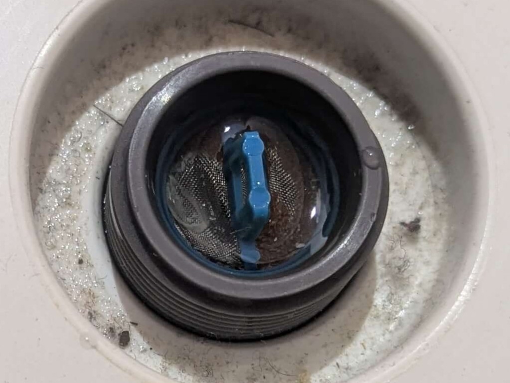 ドラム式洗濯機の給水弁フィルターに付着した鉄さびと白いカルキ（水垢）。詰まりによって水量が半減し、洗剤が溶け残って黒ワカメの温床になるメカニズムの起点。半年〜1年に1回の定期清掃が推奨。黒いワカメ発生の連鎖原因となる重要チェック箇所