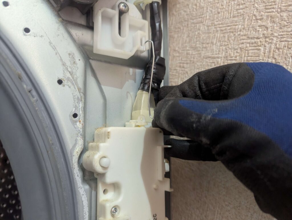 東芝 TW-160SCH ドラム式洗濯機の前面板金パネル取り外し前にドア開閉ボタンとラッチを繋ぐフックを外している状態。ラッチ・配線ケーブルも同時に取り外す。フックを外さずに板金パネルを引き抜くとラッチが破損するリスクがある