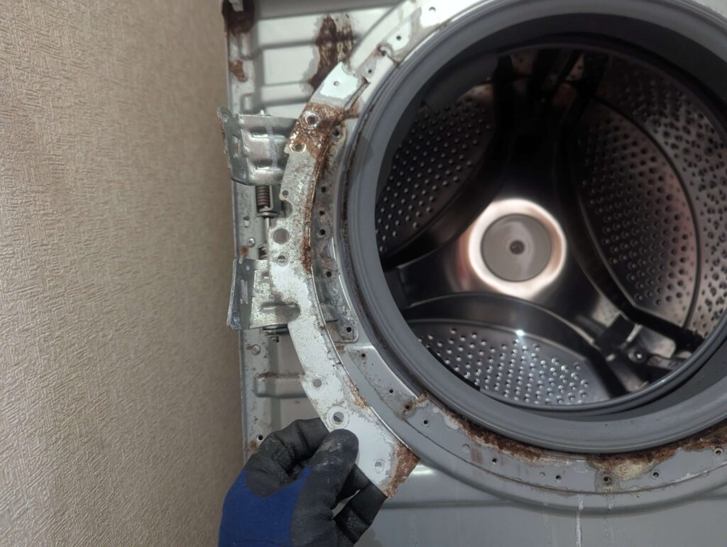 東芝 TW-160SCH ドラム式洗濯機のドアパッキン押えプレート4枚を固定ネジで外した状態。ドアパッキン本体の取り外し前に必要な手順。腐食・白粉吹きが見られる押えプレート