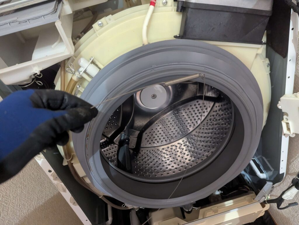 東芝 TW-160SCH ドラム式洗濯機のドアパッキン固定スプリングワイヤーを手で広げながら溝にはめ込んで取り付けている状態。工具を使わず手作業でワイヤーを広げてはめ込む再組み付け手順