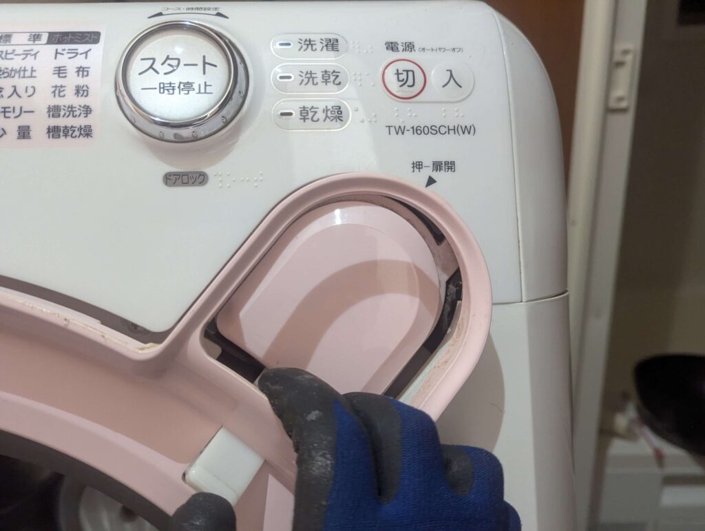 東芝 TW-160SCH ドラム式洗濯機のドア回りのカバー（ピンク色）をボタンの箇所から外している状態。差し込み爪があるため折らないよう注意が必要。外装カバー分解の手順と取り外しのコツ