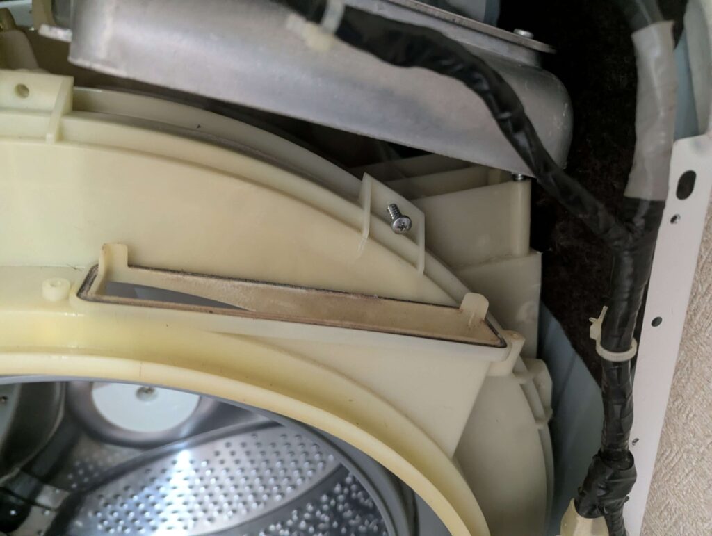 東芝 TW-160SCH ドラム式洗濯機の外槽前カバーを開けるために固定ネジを外してヒーターユニットの黒いダクトを取り外した状態。取り付けネジを機内に落とすと回収が非常に困難なため慎重な作業が必要。外槽前カバー取り外しの前提となる障害物排除工程
