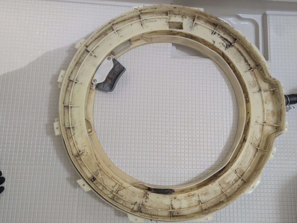 東芝 TW-160SCH ドラム式洗濯機の外槽前カバー内側の清掃前(Before)。ヘドロ化した洗剤・柔軟剤カスとカビが全面に付着した状態。歯ブラシによるブラッシング清掃前