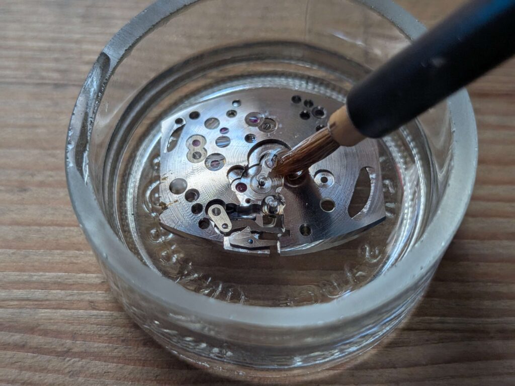 ガラス容器に入ったベンジンとハケを使い、グリュエンのムーブメント(地板)を丁寧に手作業で洗浄している様子