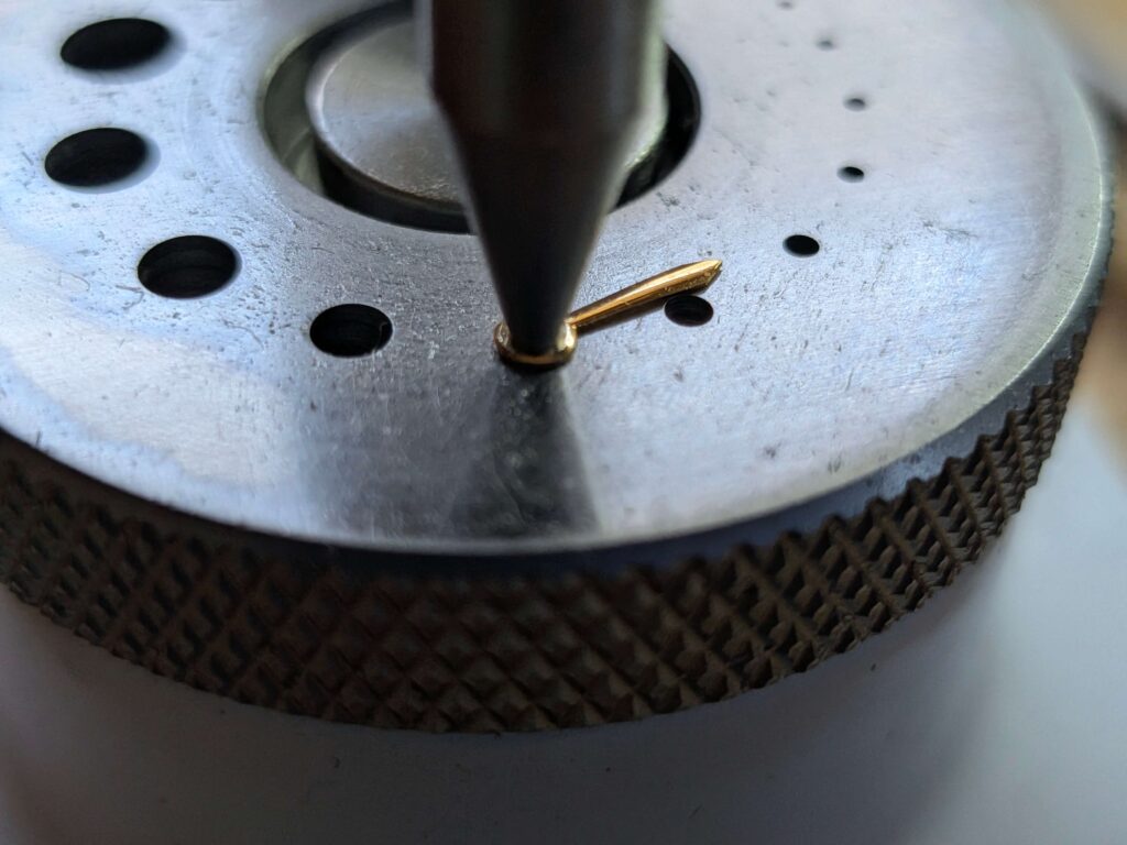 ポンス台(タガネ組み工具)を使い、ヴィンテージ時計の金色の時針の穴を微調整するためにタガネで慎重に広げている様子