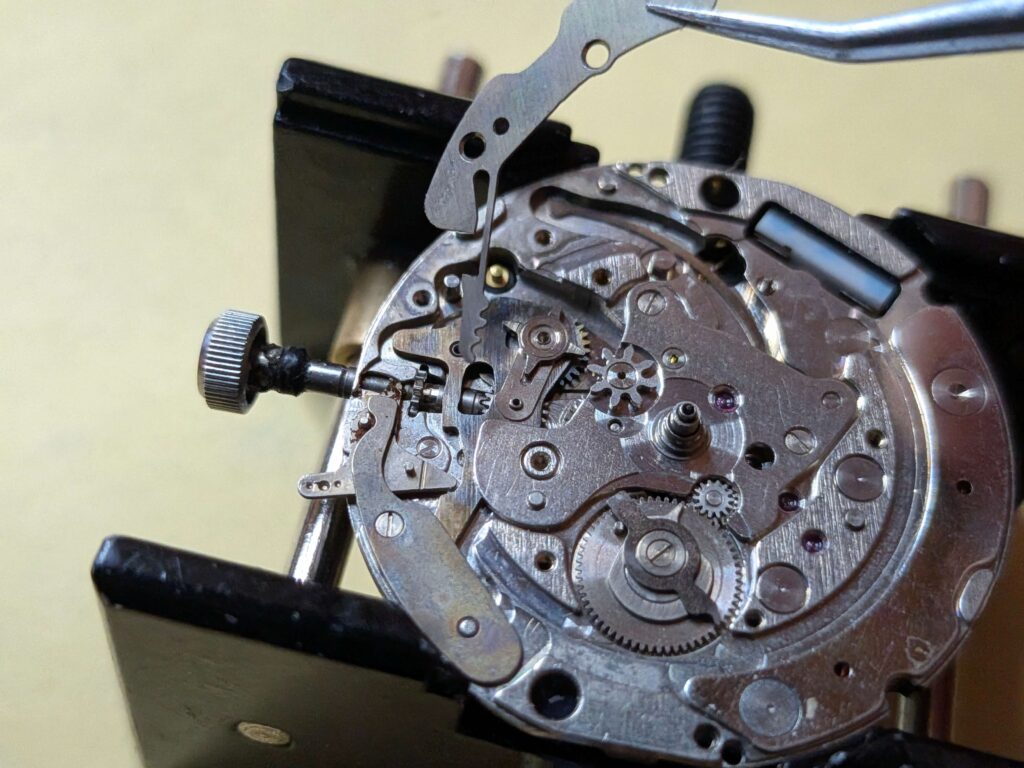 ムーブメントホルダーに固定された時計から、ピンセットで裏回りの押さえ部品を持ち上げて取り外す工程