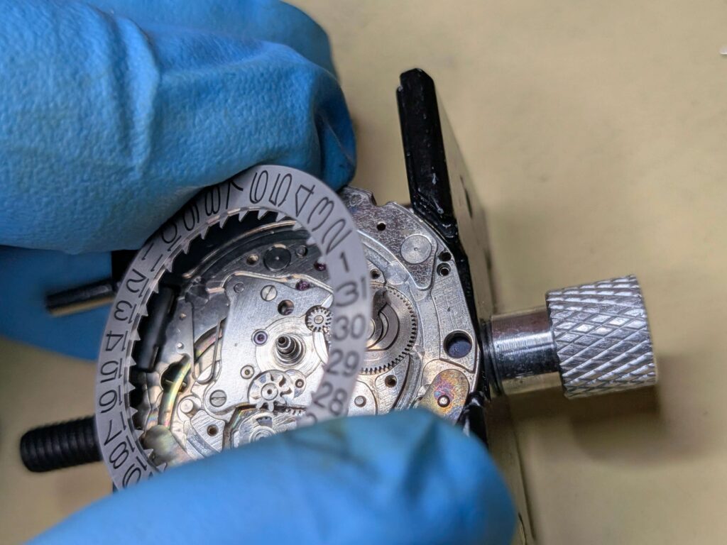 青い保護手袋を着用した手で、カレンダー機構のムーブメントから日車（日付リング）を外す作業